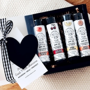 May include: A gift set featuring four Milk 'n Honey SPF 30 sunscreen lip balms in Orchard Peach, Black Cherry, Piña Colada, and Wild Orange flavors. The tubes are displayed in a black box with a heart-shaped tag and a black and white ribbon.