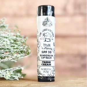 May include: A black tube of One Blessed Acre Farm Milk 'n Honey SPF 30 Sunscreen Lip Balm Fresh Cream with a white label featuring a cow and floral design. Net weight 0.15 oz (4.25g).