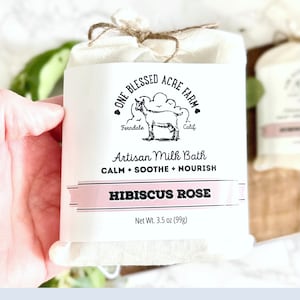 May include: A white muslin bag tied with brown twine, containing a white label with black text. The label reads "One Blessed Acre Farm", "Ferndale, Calif.", "Artisan Milk Bath", "Calm + Soothe + Nourish", "Hibiscus Rose", and "Net Wt. 3.5 oz (99g)".