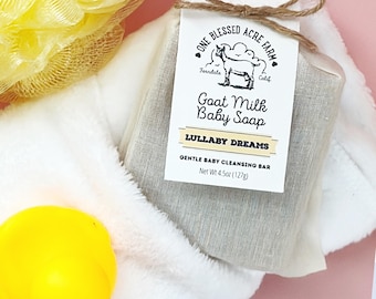 Puede incluir: Una barra de jabón para bebés de leche de cabra, envuelta en tela beige y atada con hilo, descansa sobre una superficie blanca y esponjosa. La etiqueta dice "Lullaby Dreams". También se ve un patito de goma amarillo y una esponja de baño amarilla.