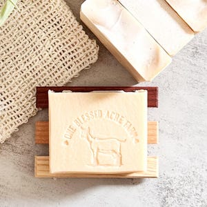 May include: A rectangular bar of off-white soap featuring an embossed goat graphic and the text "ONE BLESSED ACRE FARM" resting on a three-slat wooden soap dish, accompanied by a beige woven mesh washcloth and additional soap bars nearby.