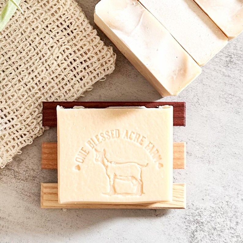 May include: A rectangular bar of off-white soap featuring an embossed goat graphic and the text "ONE BLESSED ACRE FARM" resting on a three-slat wooden soap dish, accompanied by a beige woven mesh washcloth and additional soap bars nearby.
