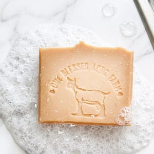May include: A rectangular bar of light brown soap with the text "ONE BLESSED ACRE FARM" and a goat silhouette. The soap is surrounded by white bubbles, suggesting a natural, handmade product. The soap appears to be sitting on a white surface.