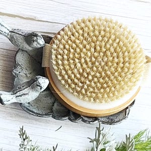 May include: A round wooden body brush with dense, light tan natural bristles and a beige elastic hand strap, resting on a decorative gray cast metal tray shaped like a leaf with two small bird figures attached to the side of the tray.