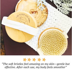 May include: Two round wooden body brushes with light-colored natural bristles and white fabric hand straps. One brush features a branded logo with the text One Blessed Acre Farm and a goat illustration. The brushes are positioned on a white cloth.
