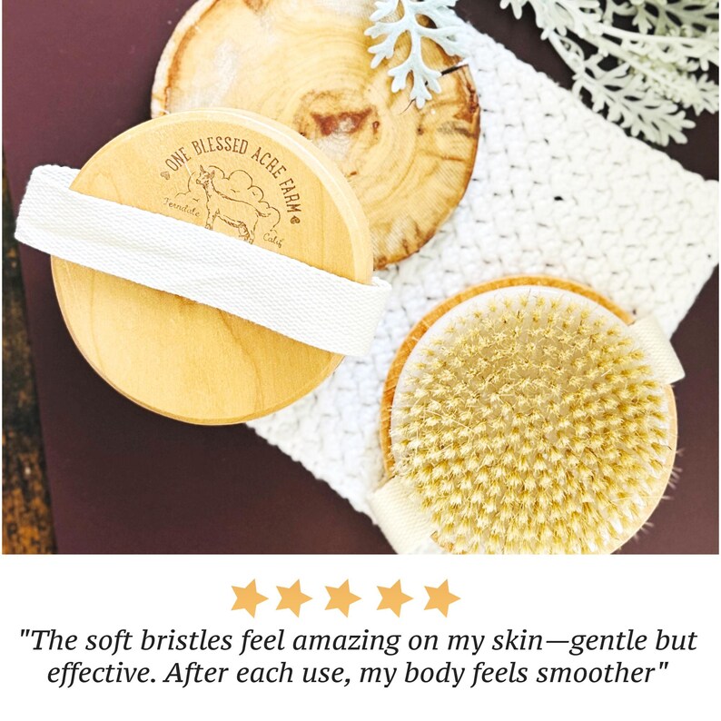 May include: Two round wooden body brushes with light-colored natural bristles and white fabric hand straps. One brush features a branded logo with the text One Blessed Acre Farm and a goat illustration. The brushes are positioned on a white cloth.