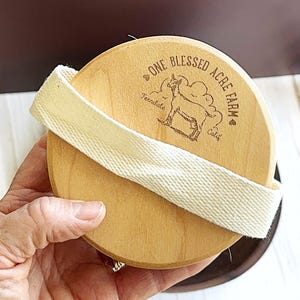 May include: A round, light-colored wooden brush featuring a cream-colored fabric strap across the center. The wood surface is branded with a graphic of a goat and the text One Blessed Acre Farm, Ferndale, Calif, with small heart icons.