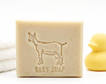 May include: A light beige bar of baby soap with a goat silhouette and the words "BABY SOAP" embossed on the front. A yellow rubber duck and a stack of white towels are in the background.
