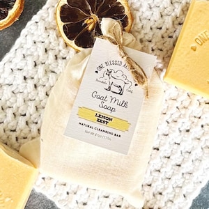May include: A white muslin bag with a label that reads "One Blessed Acre Goat Milk Soap Lemon Zest Natural Cleansing Bar Net Wt 4 oz (117g)". The bag is tied with twine and a dried lemon slice. A bar of yellow soap is visible in the background.