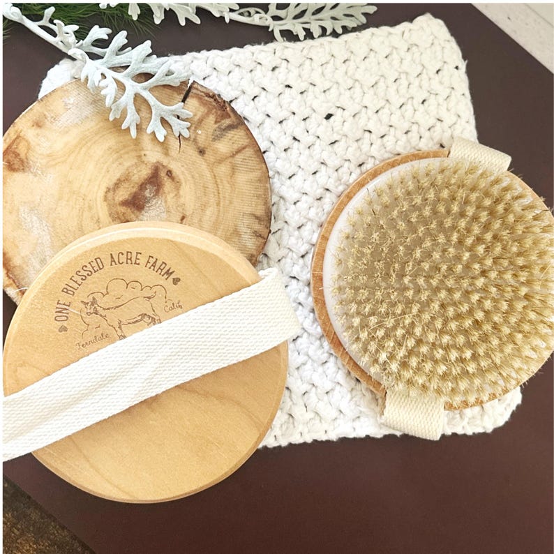May include: Two round wooden body brushes with natural bristles and cream-colored fabric hand straps. One brush shows the laser-engraved text One Blessed Acre Farm with a goat illustration. The brushes are positioned on a white crocheted cloth.