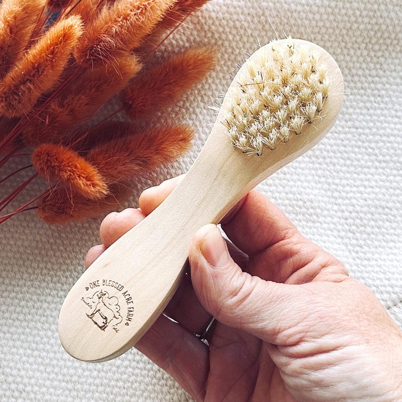 May include: A wooden facial brush with light-colored natural bristles and a smooth, light-toned wooden handle. The handle features a small, dark-inked logo that reads One Blessed Acre Farm with a graphic of a goat centered within the text.
