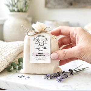 May include: A bar of goat milk soap in a beige fabric drawstring pouch with a white tag labelled One Blessed Acre Farm Goat Milk Soap Fresh Lavender Natural Cleansing Bar Net Wt 4.5oz (127g) tied with twine and decorated with lavender sprigs.