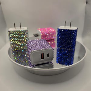 Może przedstawiać: Cztery błyszczące ładowarki USB w kolorach iryzującym, różowym, fioletowym i niebieskim. Ładowarki pokryte są małymi, błyszczącymi kryształkami. Ładowarki są prezentowane na białej, okrągłej platformie.