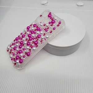 May include: Clear phone case adorned with a mix of pink and white pearls and rhinestones. The case is covered with various sizes of pearls and rhinestones, creating a textured, decorative surface. The design is arranged in a scattered pattern across the phone case.
