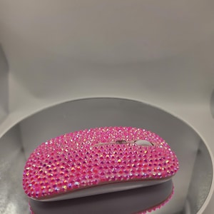 May include: A wireless computer mouse completely covered in small, sparkling pink rhinestones. The device features a white scroll wheel positioned between the two main buttons, with the entire top surface densely encrusted in reflective pink crystals.