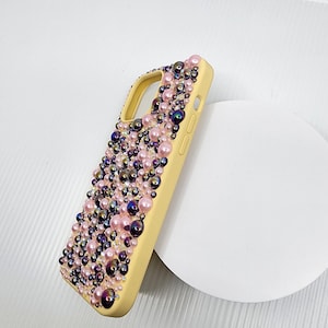May include: A yellow phone case adorned with an array of pink, purple, and iridescent beads. The case is covered in a textured pattern of various sized beads, creating a luxurious and eye-catching design. The phone case is set against a white background.