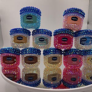 Może przedstawiać: Kolekcja słoiczków do pielęgnacji ust Vaseline, każdy ozdobiony błyszczącymi kryształkami. Słoiczki mają różne smaki, takie jak "Arctic Mint", "Wild Berry" i "Rosy Lips". Wieczka są w kolorze ciemnoniebieskim, a słoiczki ułożone są na lustrzanej powierzchni.