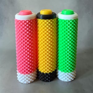May include: Three textured bicycle grips in vibrant colors. The grips are cylindrical with a diamond pattern. The colors are pink, yellow, and green, each with a contrasting top and bottom. The pink grip has a white top and bottom, the yellow has a black top and bottom, and the green has a white top and bottom.