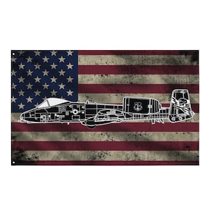 May include: A distressed American flag design featuring a black and white outline of an A-10 Thunderbolt II aircraft. The flag has red and white stripes and a blue field with white stars. The aircraft's wings, tail, and cockpit are clearly visible.