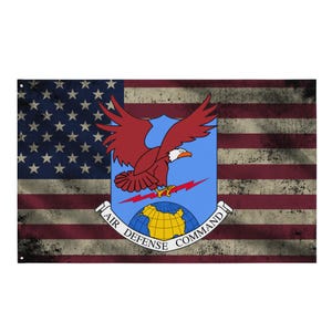 Może przedstawiać: Zniszczona flaga amerykańska z emblematem Dowództwa Obrony Powietrznej. Emblemat przedstawia czerwonego orła, niebieskie tło i napisy "Air Defense Command". Flaga ma wygląd vintage.