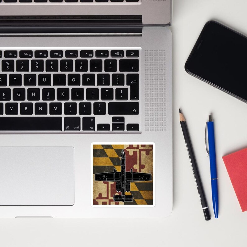 May include: A black sticker featuring a silhouette of a military aircraft, set against a background of a Maryland flag design. The sticker is placed on a laptop, with a smartphone, pens, and a notepad nearby.