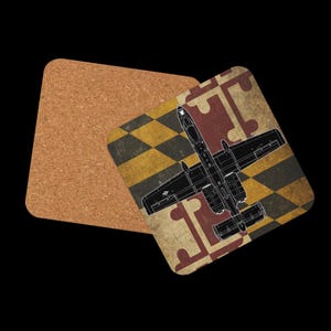 K&ouml;nnte beinhalten: Zwei quadratische Untersetzer. Einer ist aus Kork. Der andere Untersetzer hat ein Maryland-Flaggen-Design mit einer schwarz-wei&szlig;en Umrisszeichnung eines A-10 Thunderbolt II-Flugzeugs. Der Hintergrund ist schwarz.