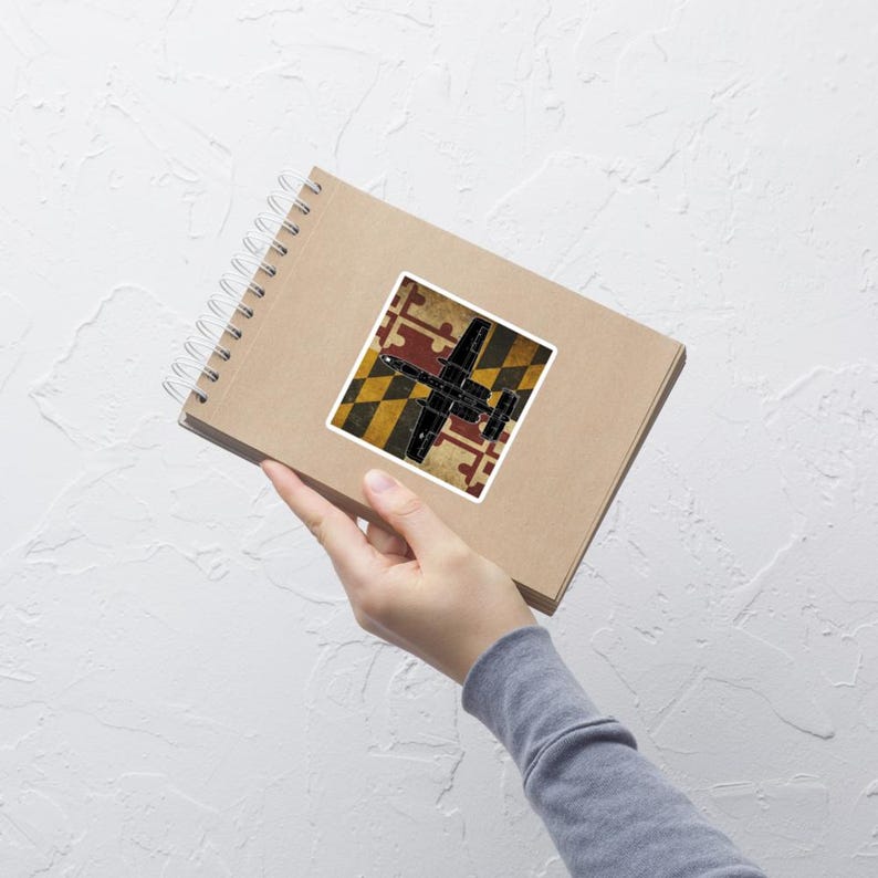 May include: A square sticker featuring a black airplane design over a Maryland flag pattern. The flag design includes red, yellow, black, and white elements. The sticker is on a spiral-bound notebook.