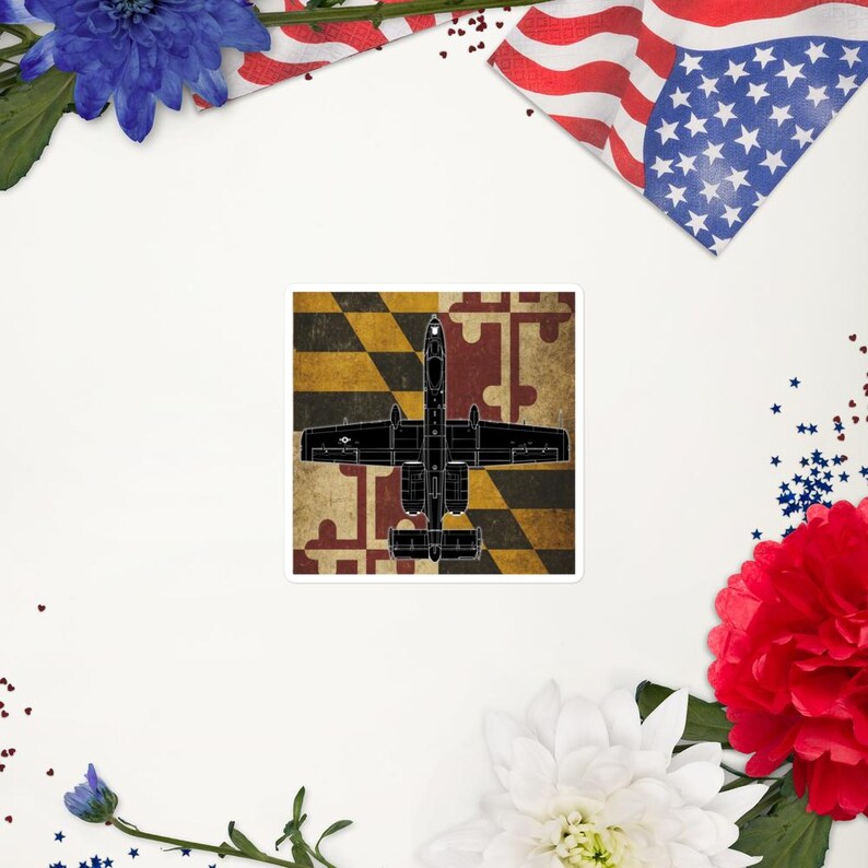 May include: A square sticker featuring a black outline of a military aircraft over a Maryland flag design. The background has a distressed, vintage look. The sticker is surrounded by patriotic decorations.