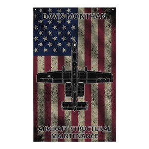 Op de afbeelding: Een verticale rechthoekige vlag met een verweerde Amerikaanse vlag als achtergrond en een zwart silhouet van een A-10 vliegtuig in het midden. De tekst DAVIS MONTHAN staat bovenaan en AIRCRAFT STRUCTURAL MAINTENANCE onderaan in het wit.