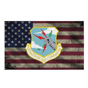 Pode incluir: Uma bandeira americana desgastada com o emblema do Comando Aéreo Estratégico. A bandeira apresenta listras vermelhas e brancas e um campo azul com estrelas brancas. O emblema inclui um escudo com fundo azul celeste, um braço prateado blindado segurando raios vermelhos e o texto "STRATEGIC AIR COMMAND".
