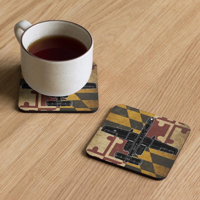 K&ouml;nnte beinhalten: Zwei quadratische Untersetzer mit einem Maryland-Flaggen-Design und einer schwarzen Flugzeuggrafik. Ein Untersetzer hat eine wei&szlig;e Tasse mit Tee darauf. Die Untersetzer befinden sich auf einer hellbraunen Holzoberfl&auml;che.