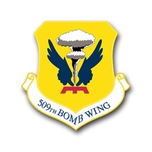 May include: A yellow shield emblem with a white banner at the bottom. The emblem features a stylized mushroom cloud, blue wings, and a red base. The banner reads "509th BOMB WING".
