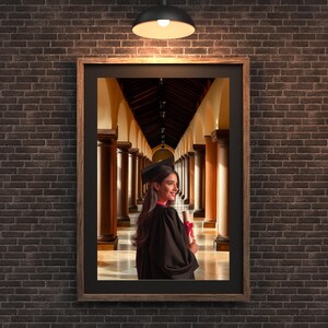 May include: A framed photograph of a graduate in a black cap and gown, holding a diploma with a red ribbon. The image is set against a backdrop of a long hallway with columns and arches, creating a sense of depth and perspective. The frame is brown, and the background is a brick wall.