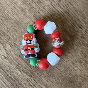 Könnte beinhalten: Ein weihnachtliches Armband mit einem Nussknacker-Anhänger, roten und weißen Perlen und grünen Akzenten. Das Armband besteht aus Perlen in verschiedenen Formen und Größen mit einem festlichen Design.