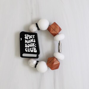 Puede incluir: Una pulsera con un dije de libro negro con el texto "Spicy Moms Book Club". La pulsera incluye cuentas geométricas blancas y color cobre, y cuentas redondas blancas. Un cierre plateado asegura la pulsera.
