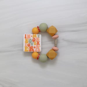 Könnte beinhalten: Ein farbenfrohes Armband mit dem Text "IN MY BOOK LOVER ERA". Es zeigt eine Mischung aus runden und geometrischen Perlen in Orange, Grün und Gelb. Die Perlen sind mit einem silbernen Verschluss aufgereiht.