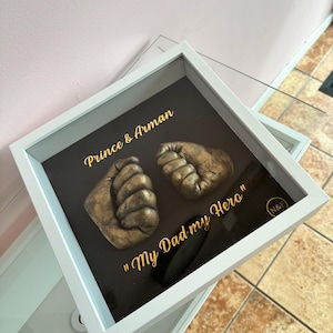 May include: A white shadow box frame containing two gold-coloured baby hand casts mounted on a dark background. The text "Prince & Arman" and "My Dad my Hero" is printed in gold lettering, with a small circular "N&F" logo in the bottom corner.