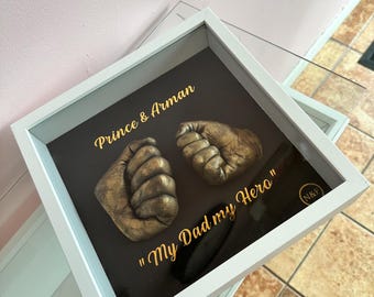 May include: A white shadow box frame containing two gold-colored baby hand casts mounted on a dark background. The text "Prince & Arman" and "My Dad my Hero" is printed in gold lettering, with a small circular "N&F" logo in the bottom corner.