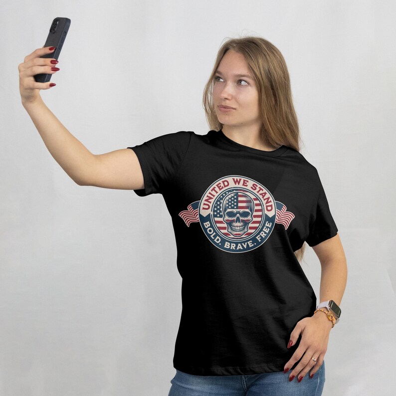 Puede incluir: Camiseta negra con un dise&ntilde;o circular que presenta una calavera que incorpora la bandera estadounidense. El dise&ntilde;o est&aacute; rodeado por las palabras "United We Stand, Bold, Brave, Free" y dos banderas americanas.