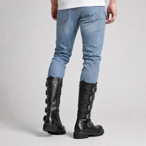 May include: Light blue denim jeans paired with black mid-calf boots. The boots feature a side zipper and five horizontal straps across the front, each secured with a silver-toned metal buckle. The jeans have a classic five-pocket design.