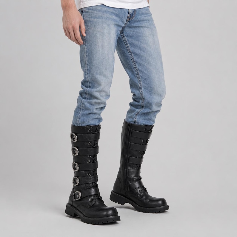 May include: Black mid-calf boots featuring five horizontal straps with silver-toned metal buckles and eyelets, paired with light blue denim jeans. The boots have a rounded toe, a thick lug sole, and a lace-up front design with a side zipper closure.