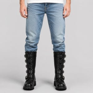 May include: Light blue denim jeans paired with black mid-calf boots. The boots feature a lace-up front design with five horizontal straps across the shaft, each secured by a circular metal buckle, and a thick, lugged rubber sole for traction.