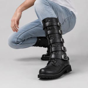 May include: A pair of black mid-calf boots featuring a lace-up front and five horizontal straps with silver-toned skull-shaped buckles. The boots have a rounded toe, a thick lug sole, and a smooth finish, extending to the mid-calf area.
