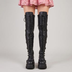 May include: Black over-the-knee boots featuring a full-length front lace-up closure with metal eyelets, multiple horizontal buckled straps along the sides, and a thick platform sole. The boots extend above the knee and have a smooth matte finish.