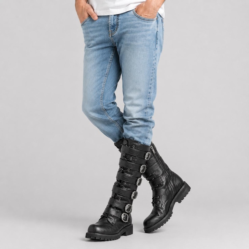 May include: Light blue denim jeans paired with black leather mid-calf boots. The boots feature a lace-up front, a side zipper, and five horizontal straps across the front, each secured with a silver-toned metal buckle and a rounded toe design.