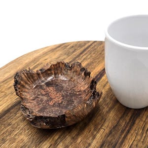 May include: A white ceramic mug with a curved handle sits on a wooden surface next to a small, irregularly shaped wooden bowl. The bowl has a dark brown, textured appearance. The wooden surface has a natural grain pattern.
