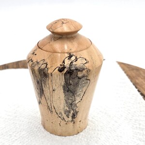 May include: A handcrafted wooden urn with a lid. The urn is light brown with dark gray and black veining patterns. The urn has a rounded body and a small, rounded lid. The wood grain is visible throughout the piece.