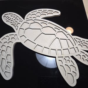 May include: A light-colored wooden sea turtle cutout with intricate details. The turtle is positioned on a black surface, showcasing its segmented shell and flipper design. The cutout is a decorative item.