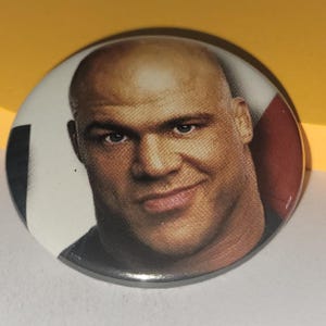 Puede incluir: Un primer plano de un botón que presenta a un hombre con la cabeza rapada y una expresión seria. El botón es redondo y tiene un borde plateado.