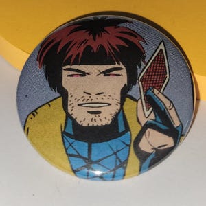 Può includere: Bottone rotondo con un'illustrazione a fumetti di un personaggio con i capelli rossi, che tiene in mano una carta da gioco. Il personaggio indossa un cappotto giallo e una camicia blu. Lo sfondo è un motivo a pois blu chiaro.